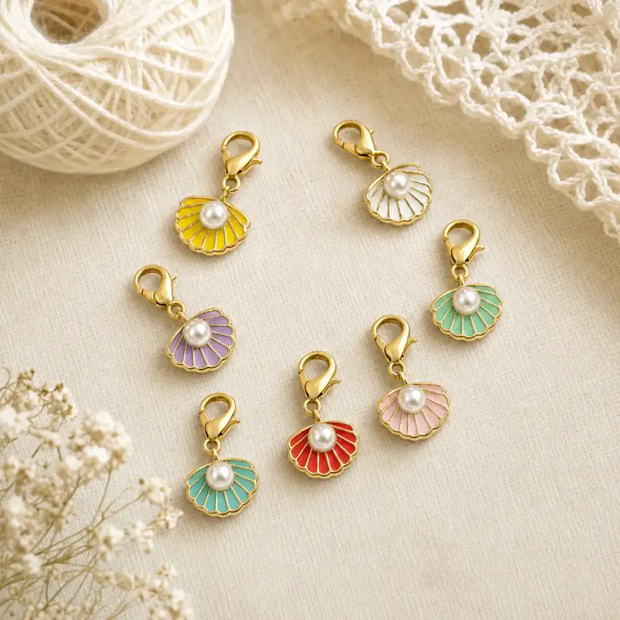 Seven pearl shell charm stitch markers arranged on cream linen beside a ball of ivory yarn